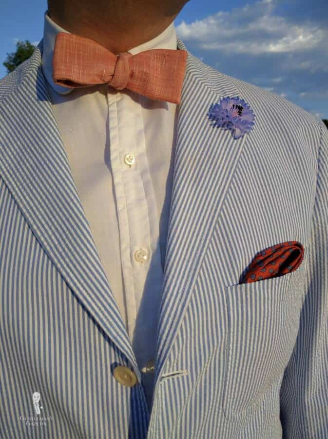 Camisa branca com terno seersucker com bolso de patch e lenço de bolso laranja e azul, gravata borboleta e lapela de Fort Belvedere
