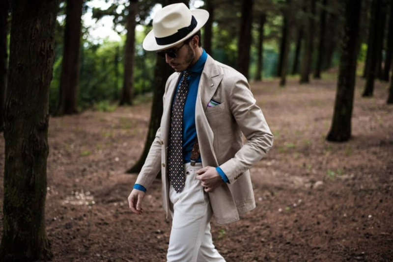 Chapeau panama, veste sable, chemise bleue, cravate en soie garance imprimée et pantalon blanc cassé