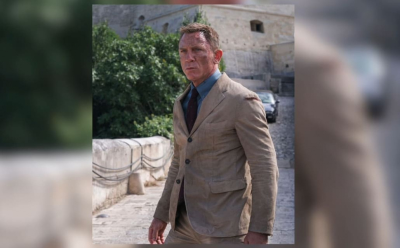 Daniel Craig jako James Bond ve filmu No Time To Die, na sobě pískový manšestrový nestrukturovaný oblek, chrpově modrou košili a kaštanovou hedvábnou kravatu s opakujícím se vzorem modrých dlaždic