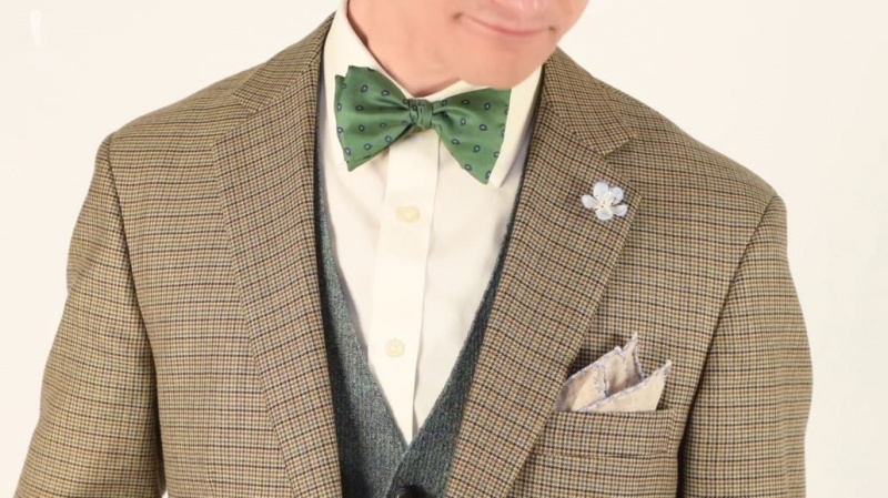Veste pied-de-poule associée à un gilet gris, une chemise blanche, une pochette en lin marron clair, une boutonnière bleu clair et un nœud papillon vert.