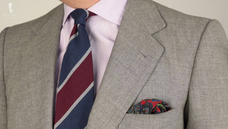 Une cravate grenadine en cachemire et laine en bleu foncé, bordeaux, rayures gris clair de Fort Belvedere, associée à un costume gris clair, une chemise lavande et une pochette à motifs