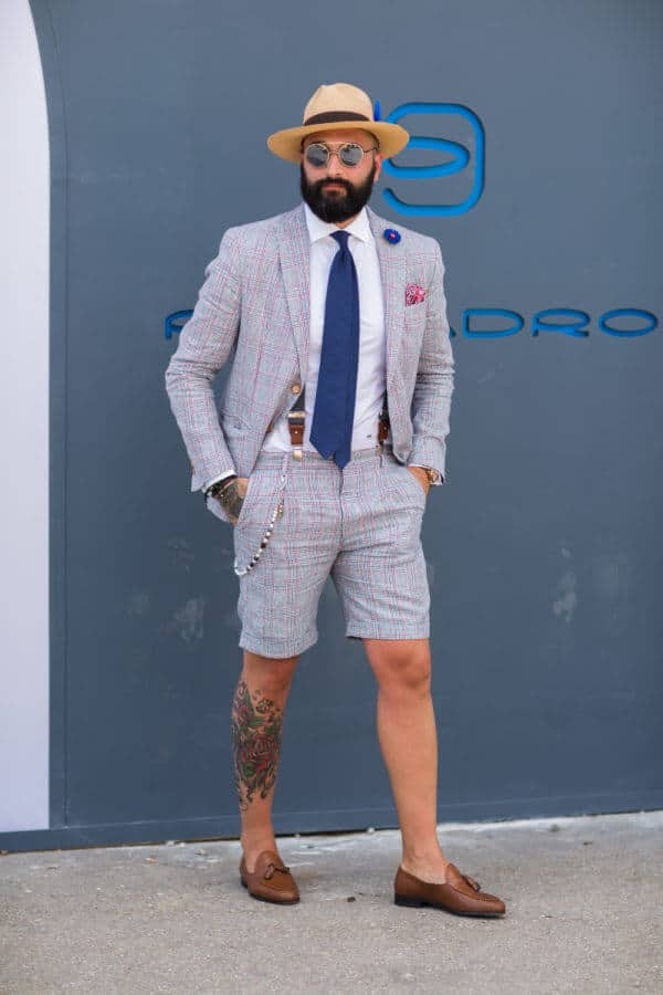 Le tailleur short avec cravate bleue, bretelles, boutonnière, pochette, chapeau, lunettes de soleil et mocassins à glands