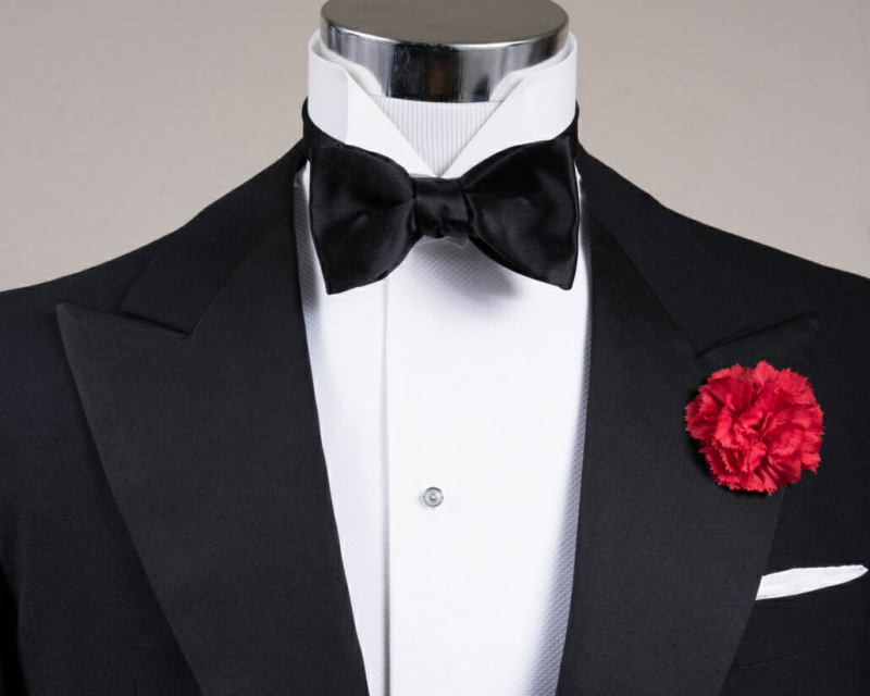 Ensemble Black Tie accessoirisé avec un nœud papillon en satin de soie noir, une pochette de costume blanche et une boutonnière à œillets rouges de Fort Belvedere.