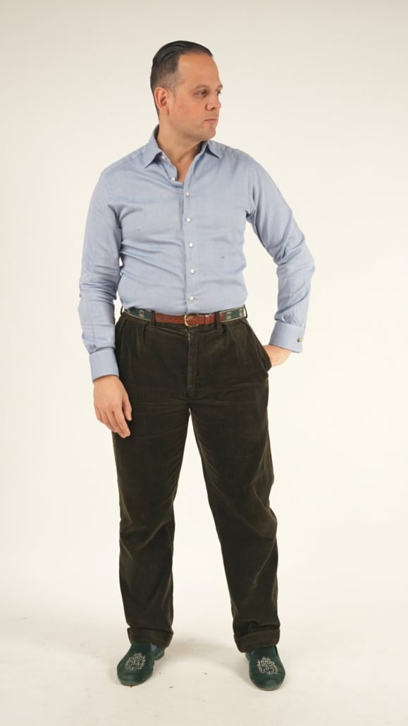 Raphael má na sobě košili ušitou na míru od 100 rukou v modrošedé barvě, tmavě hnědé manšestrové kalhoty, hnědý pásek a zelené sametové pantofle Albert s logem Fort Belvedere.