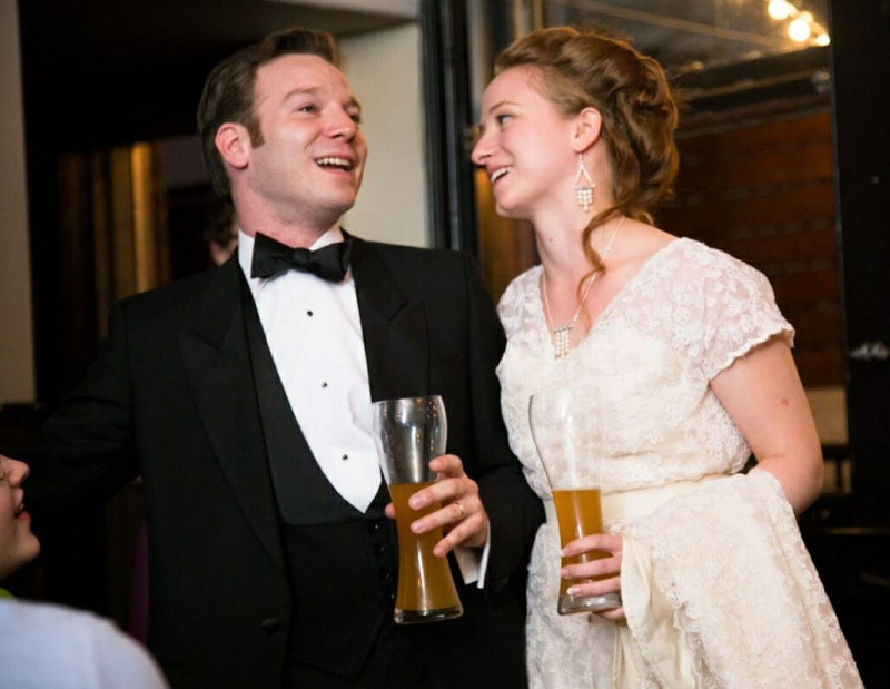 Smoking noir avec gilet de soirée en V profond, chemise blanche avec 3 boutons de chemise, nœud papillon noir à côté de la mariée, tous deux buvant de la bière de blé.