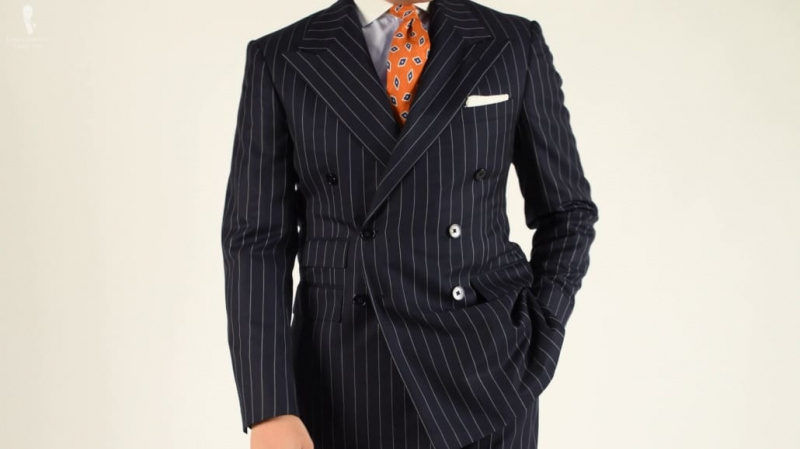 Um terno de risca de giz azul marinho com abotoamento duplo combinado com um lenço de bolso branco e gravata com estampa laranja.