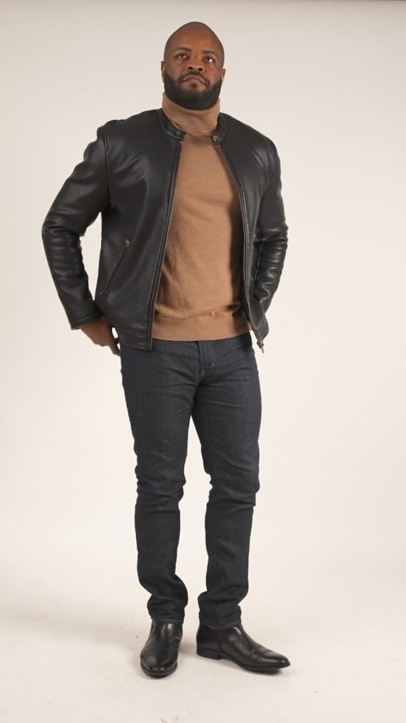 Kyle porte une veste en cuir de moto noire, un sweat-shirt à col roulé beige, un jean en denim et des bottes noires.