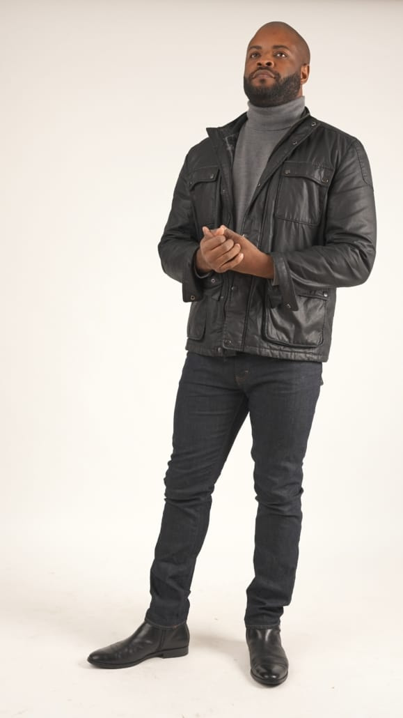 Кајл носи црну јакну од воштаног памука, сиви џемпер и тексас фармерке.