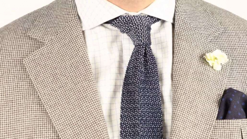 Une pochette en étamine de laine bleu marine à pois bleus associée à une veste pied-de-poule, une chemise à carreaux, une boutonnière œillet blanc et une cravate en maille bicolore marine et bleu clair.