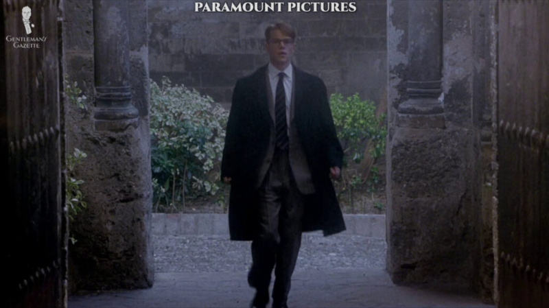 Tom Ripley porte un costume à carreaux Prince de Galles avec une chemise blanche à rayures et un pardessus foncé.