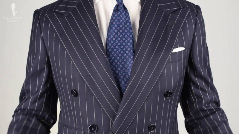 Tmavě modrý pruhovaný oblek s tužkou, bílou košilí, hedvábnou kravatou z madder se vzorem Macclesfield a bílým kapesníčkem.