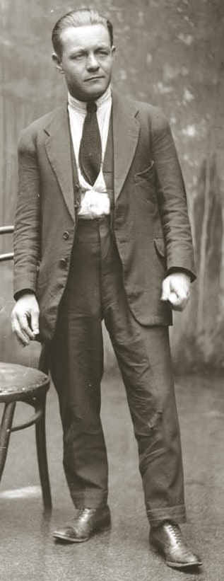Černobílá fotografie muže v denním obleku s dlouhou sukní a kalhotami s vysokým pasem.