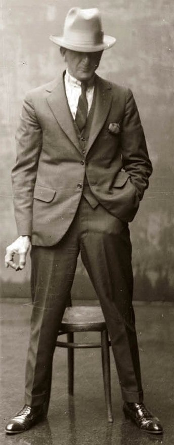 スリーピースのスーツを着てフェドーラ帽を軽快に顔の上に下ろした男性の白黒写真。