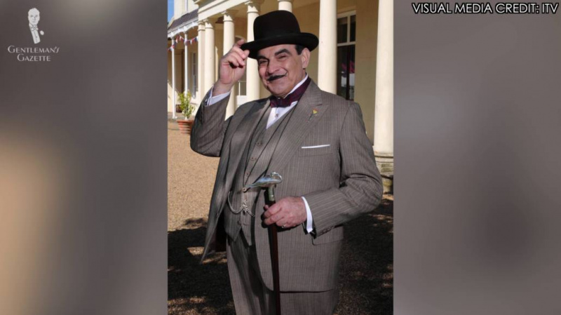 Hercule Poirot ruskeassa kolmiosaisessa raitapuvussa ja hatussa sekä keppi mukana