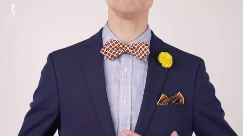 Престон носи тамноплаво одело са одштампаном лептир машном, квадратни џеп са нијансама злата и жуту дугме од маслачка.