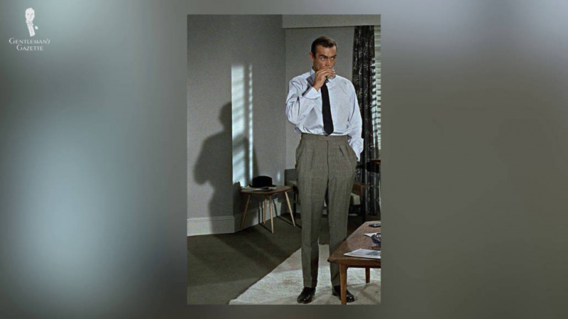 Човек који стоји обучен у светлоплаву кошуљу и црну кравату упарен са високим панталонама.