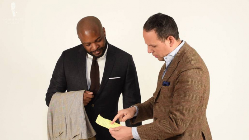 Un tailleur recevant les demandes de personnalisation du client. Kyle et Raphael se faisant passer pour un tailleur et un client.