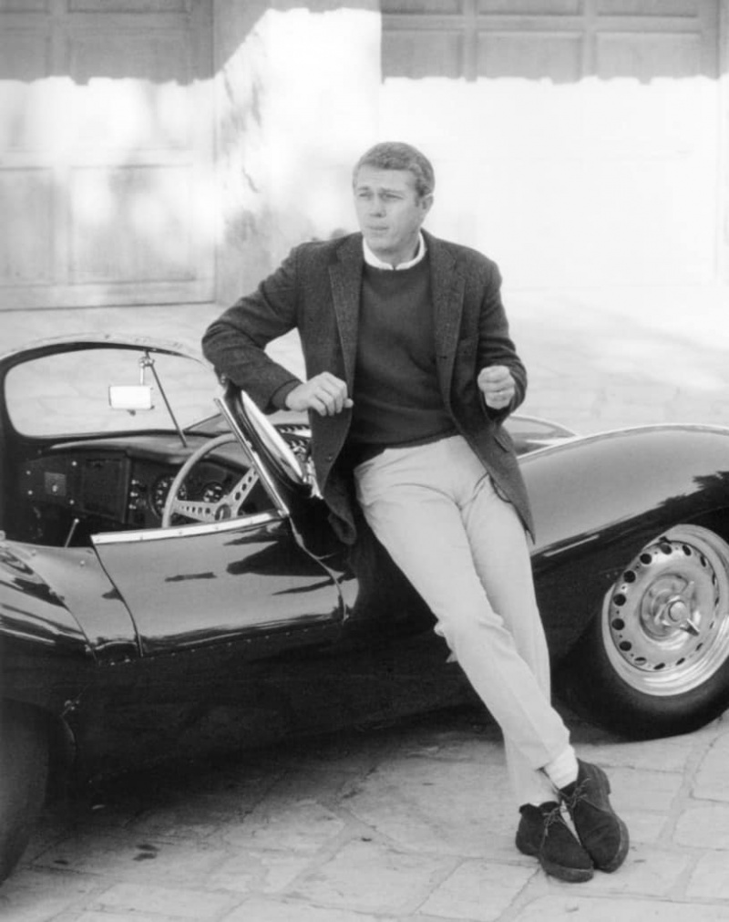 Steve McQueen porte un pantalon kaki avec des chaussettes blanches, des bottes chukka, une chemise à col rond, un pull et un manteau.