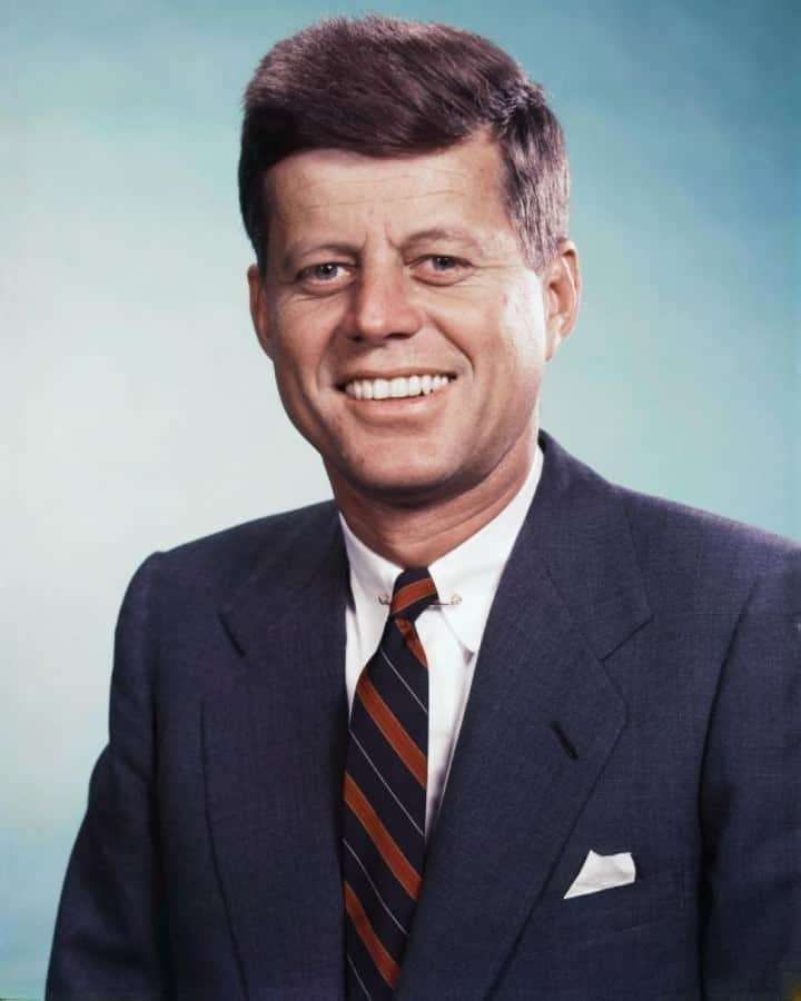 John F Kennedy souriant sur cette photo dans une veste bleu marine associée à une cravate à rayures et une pochette en lin blanc