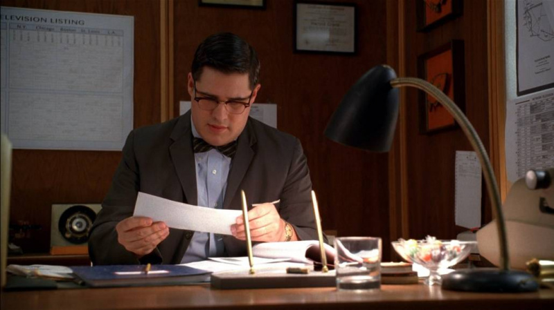 Harry Crane sentado em sua mesa e olhando para uma carta. Ele está vestindo um paletó desabotoado, uma gravata borboleta estampada e uma camisa social