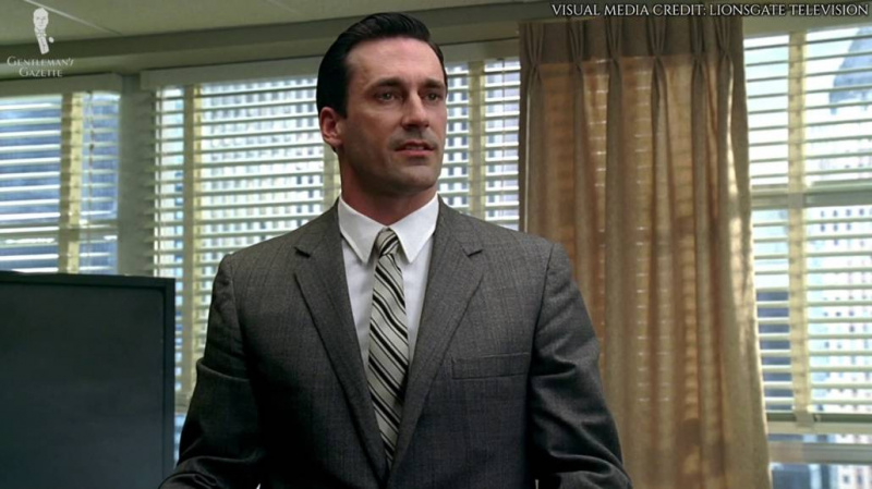 Don Draper dans un costume gris clair, une chemise blanche à col pointu et une cravate à rayures.