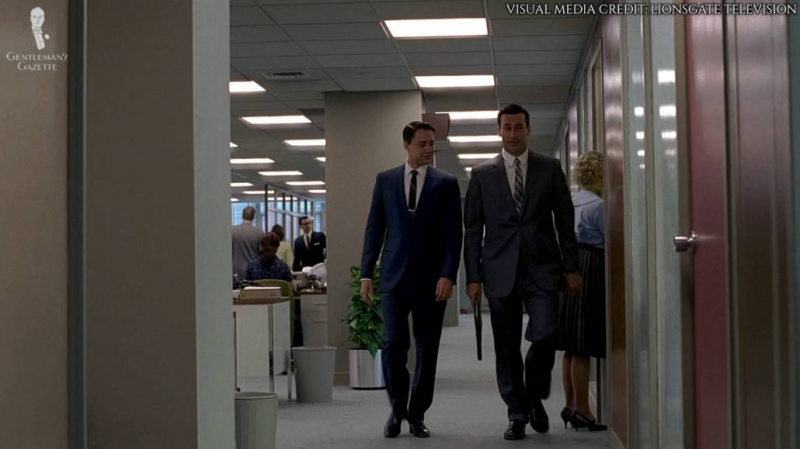 Don Draper e Peter Campbell andando pelos corredores. Ambos estão vestindo ternos de negócios.