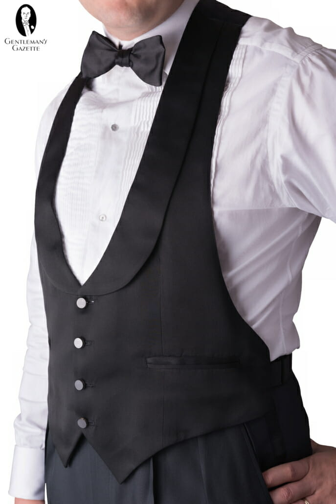 Ovaj prsluk s crnom kravatom je model bez leđa i ima četiri polirana gumba, tijelo od mat vune s okrenutim reverima i dva