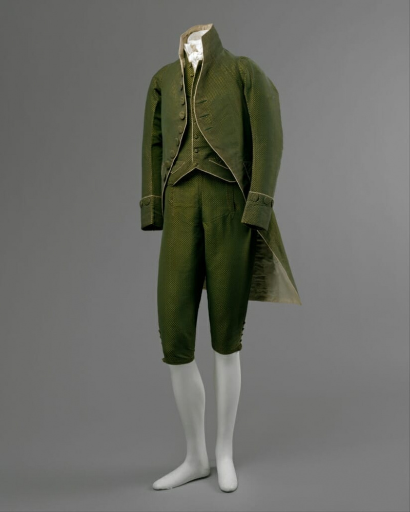 Cette tenue de c. 1790 comprend un manteau à manches étroites et une jupe découpée, un gilet court et une paire de culottes. Une certaine opulence demeure dans son tissu luxueux.