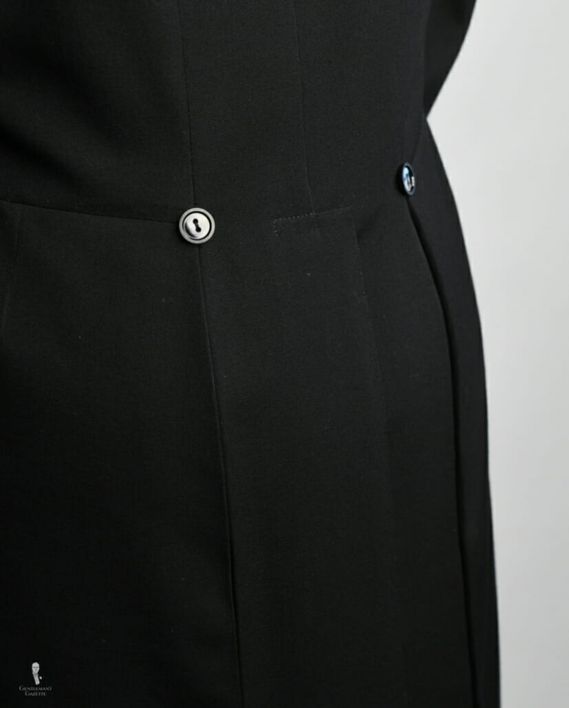 Costas de fraque com costuras verticais e horizontais, abertura central do jarrete e dois botões nas costas, uma relíquia de quando o casaco de montaria era abotoado na parte de trás