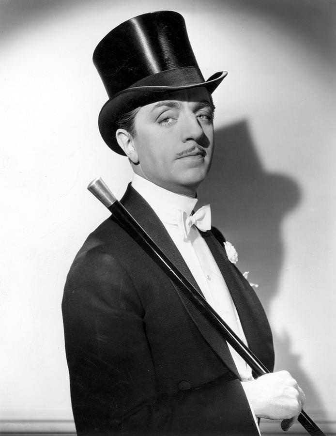 William Powell en queue de pie à col châle, boutonnière, col cassé, avec canne et chapeau haut de forme en soie